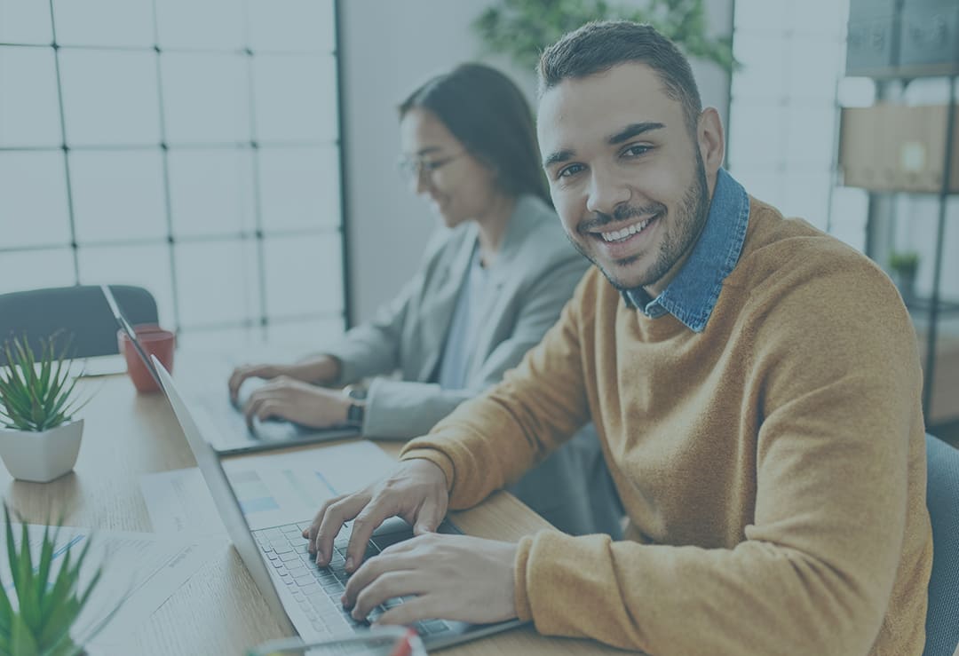 smiling male and female employees working on a laptop beside a glass window